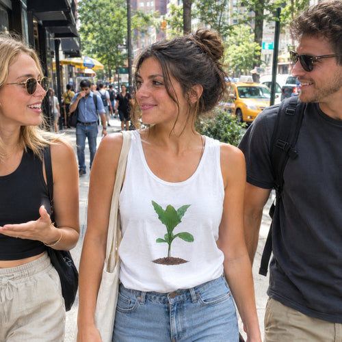 A woman walking on a city sidewalk wearing a white tank top with a green seedling graphic, paired with light blue jeans and carrying a beige shoulder bag. She is surrounded by two men in an urban street setting.